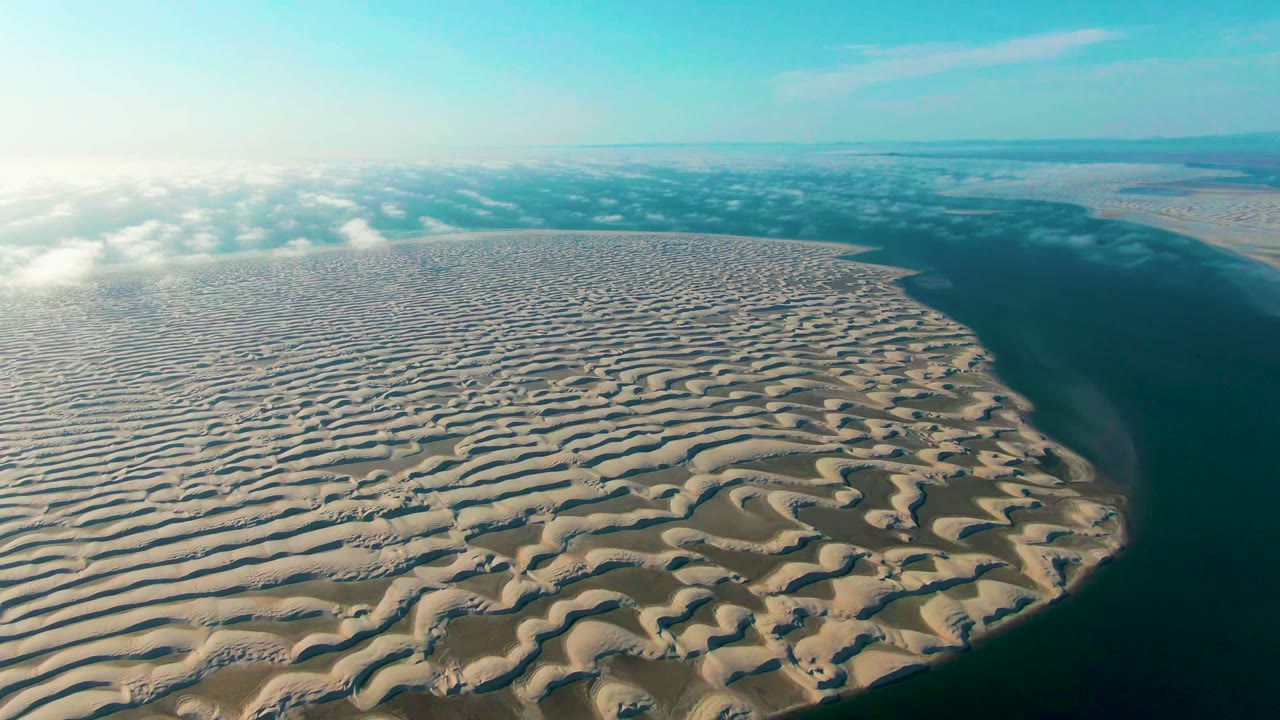 Aerial frame from Guerrero Negro showing a broad beige dune field, darker blue-green water running along the right side, a distant flat horizon, and no visible people.