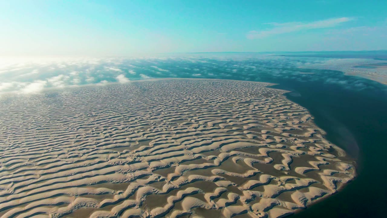Opening aerial frame over rippled sand dunes in Guerrero Negro, Baja California Sur, with dark water along the right edge, low clouds on the horizon, and no visible people.