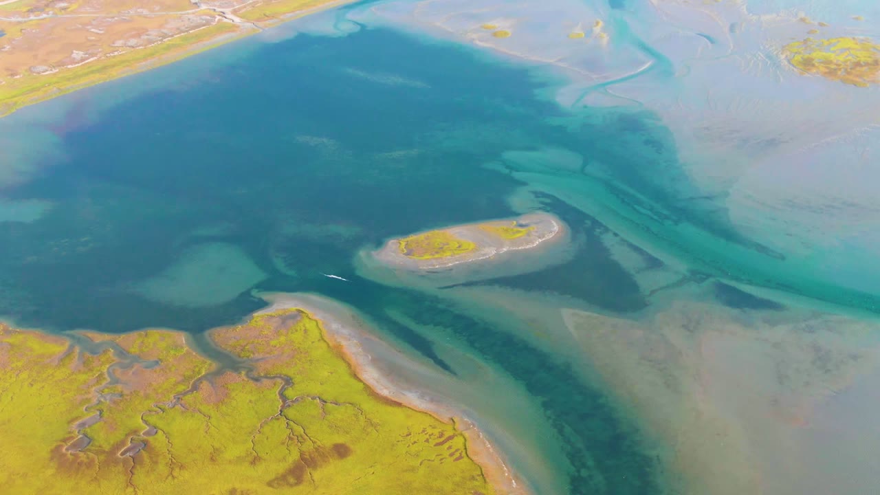 A high aerial frame over Guerrero Negro wetlands shows dark blue water, turquoise shallows, yellow-brown marsh, a small island-like sandbar, and no visible people.