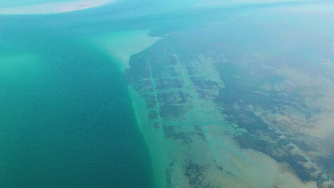 Aerial frame over Guerrero Negro, Mexico, showing turquoise ocean on the left, pale shallows along the shore, gridded coastal land on the right, and no visible people.