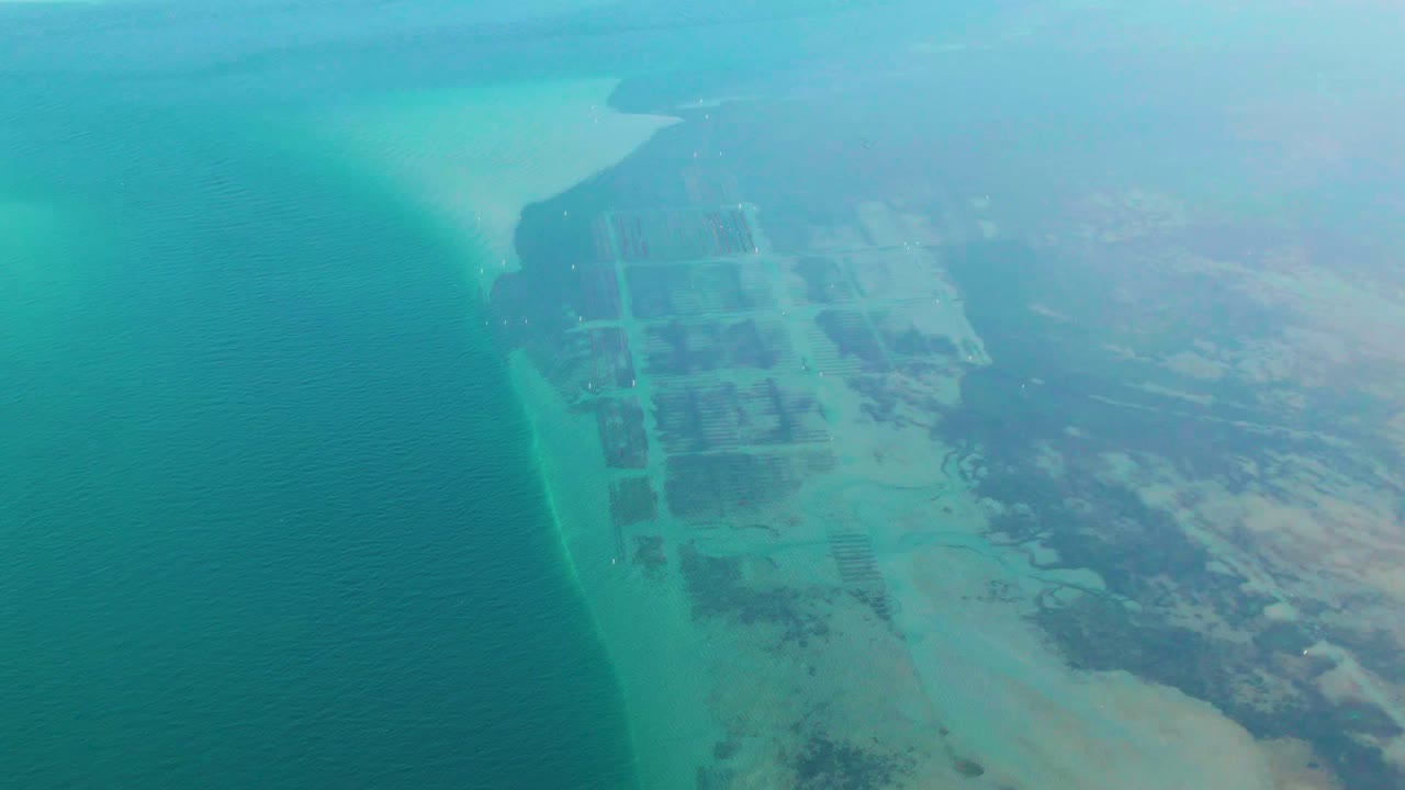 Aerial view of Guerrero Negro coastline with open turquoise water, a thin surf or shoal line, structured land parcels, distant haze, and no visible people.