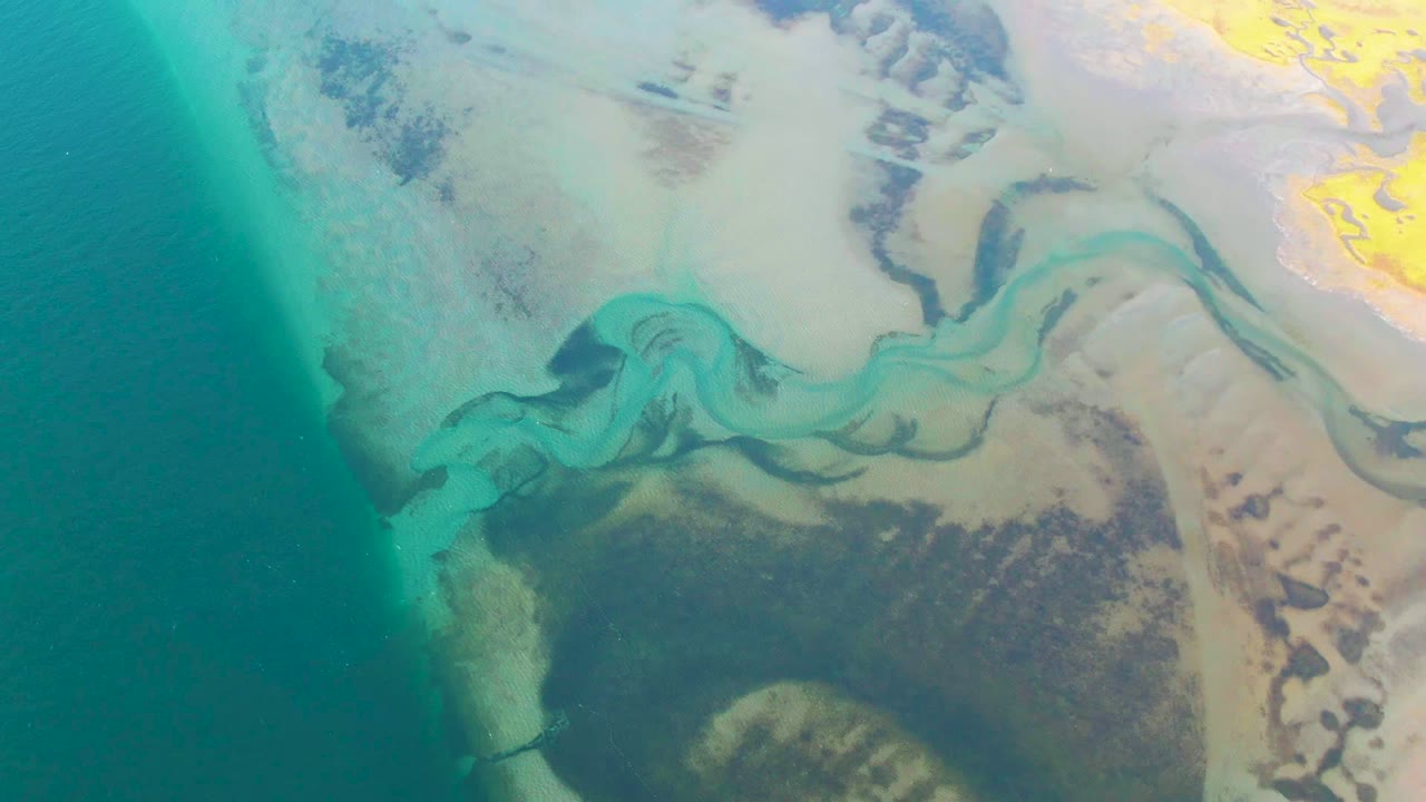 Overhead view in Guerrero Negro, Baja California Sur, with turquoise water at left, pale sandbars, dark mudflat areas, and a curving stream channel, no visible people.