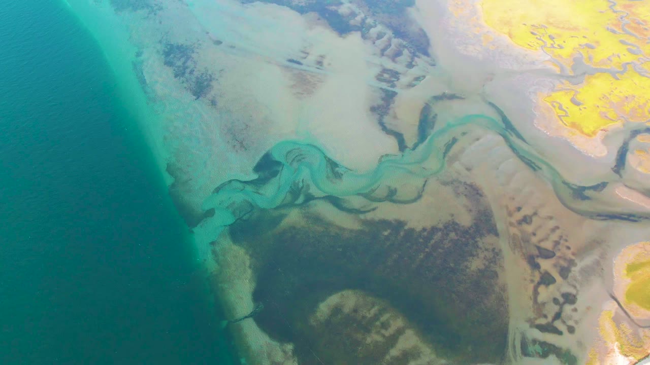 Aerial view over Guerrero Negro coastal flats where turquoise ocean borders pale sand, a sinuous estuary channel, dark wetland patches, and yellow mudflat areas, no visible people.