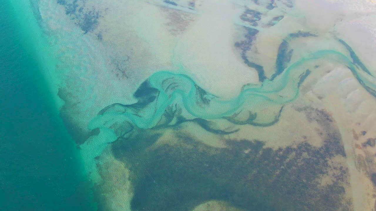 Overhead view in Guerrero Negro, Baja California Sur of a turquoise winding channel crossing pale sandy shallows, with darker submerged patches and no visible people.