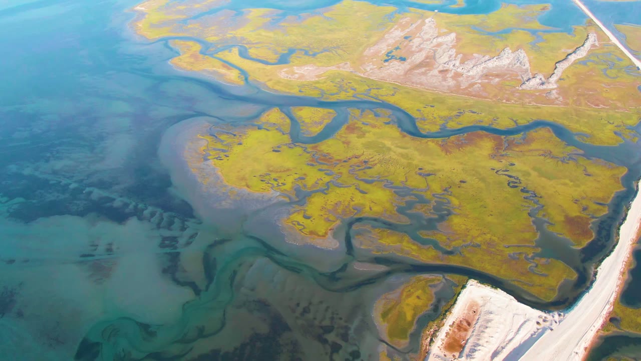 Aerial view over Guerrero Negro wetland channels, blue-green shallow water, yellow marsh vegetation, pale shoreline, a straight road at right, distant low hills, and no visible people.