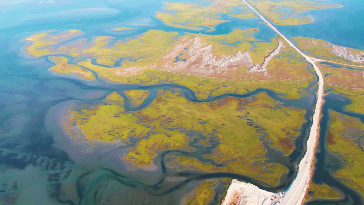 Aerial frame from Guerrero Negro, Baja California Sur, showing a pale road along the right side, green wetland patches, blue shallow water, and no visible people.