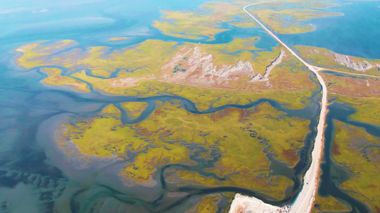 Aerial frame from Guerrero Negro, Baja California Sur, showing the pale road curving toward the top right, dark waterways through green marsh, blue water flats, and no visible people.