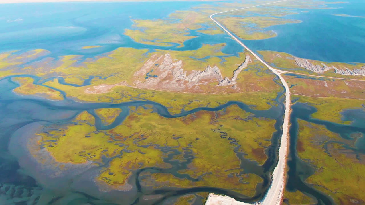 Aerial frame from Guerrero Negro, Baja California Sur showing a pale road cutting through green-yellow marsh, winding dark channels, blue water, and no visible people.