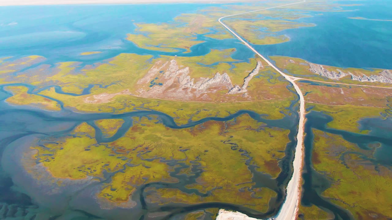 At 0:02, the aerial composition shows the long road edging past marsh islands, dark sinuous waterways, open blue water, and no visible people in Guerrero Negro.
