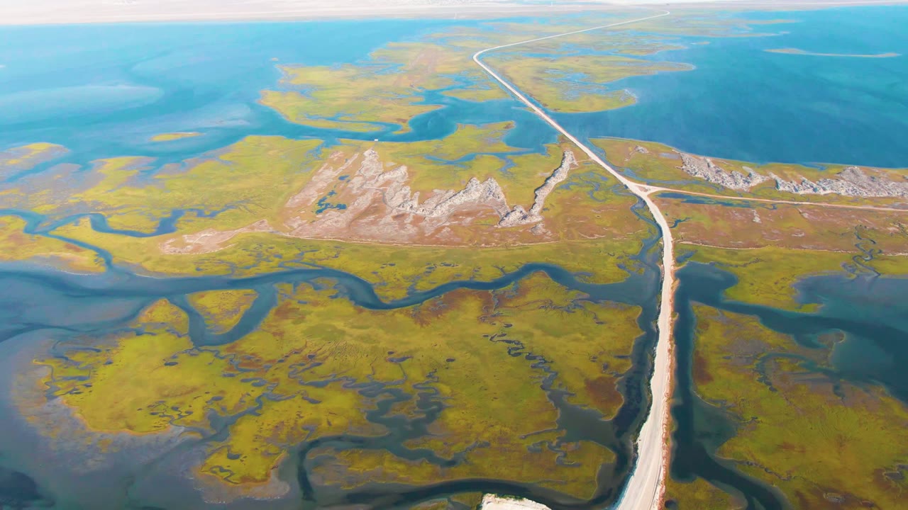 A high aerial frame over Guerrero Negro shows a pale road cutting through green marsh, blue water channels, and rocky low terrain, with no visible people.