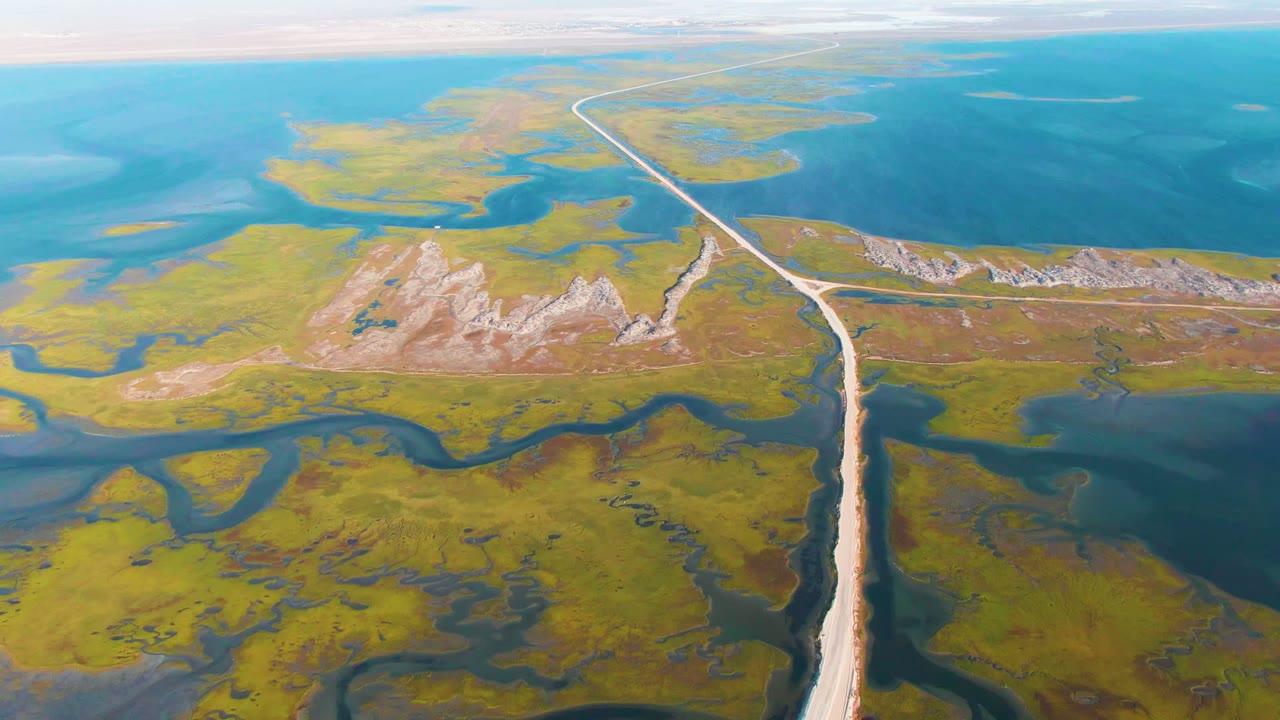 Aerial frame over Guerrero Negro wetlands with a pale dirt road rising from the lower edge toward blue water, green marsh channels, rocky light ground, and no visible people.