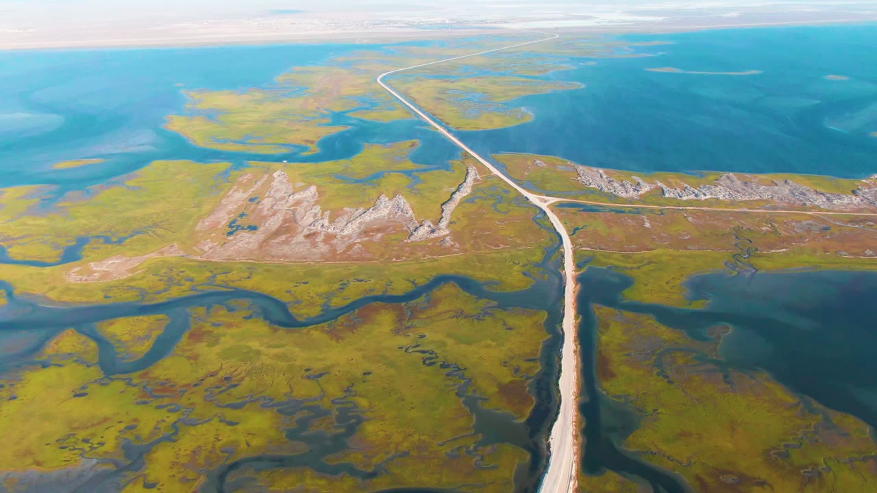 High aerial frame over Guerrero Negro showing the road crossing green wetland terrain, pale rocky patches near the center, broad blue water in the background, and no visible people.