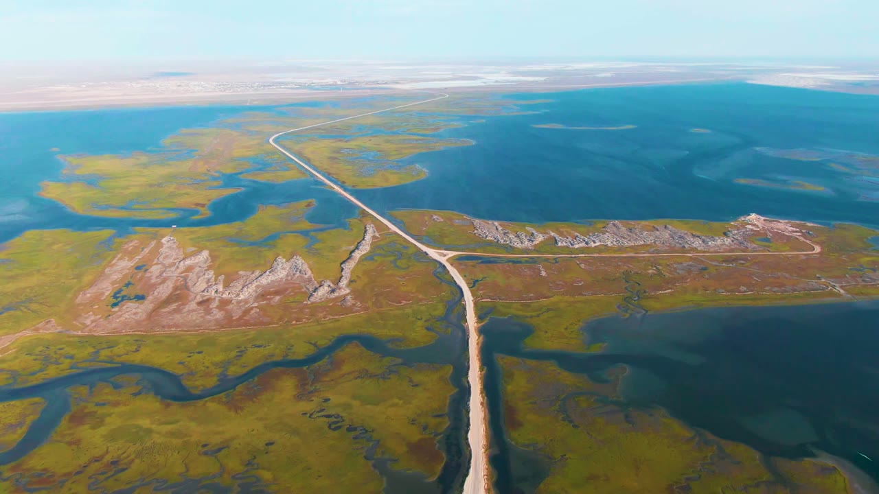 Opening aerial frame over Guerrero Negro, Baja California Sur, with a pale dirt road running from the lower edge through green wetland flats toward wide blue water and no visible people.