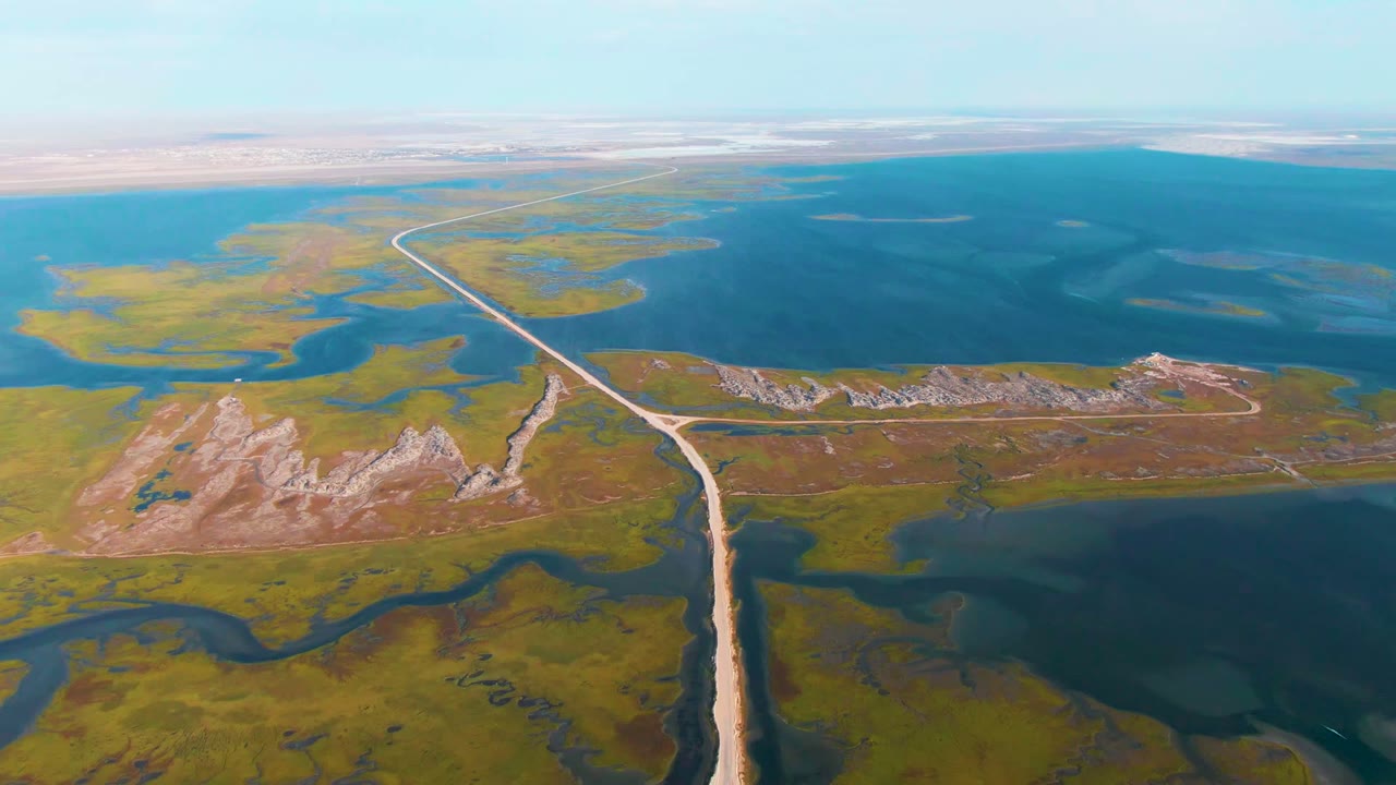 Mid-clip aerial view over Guerrero Negro with the dirt road angling across marshy green terrain, dark water channels in the foreground, broad blue water beyond, and no visible people.
