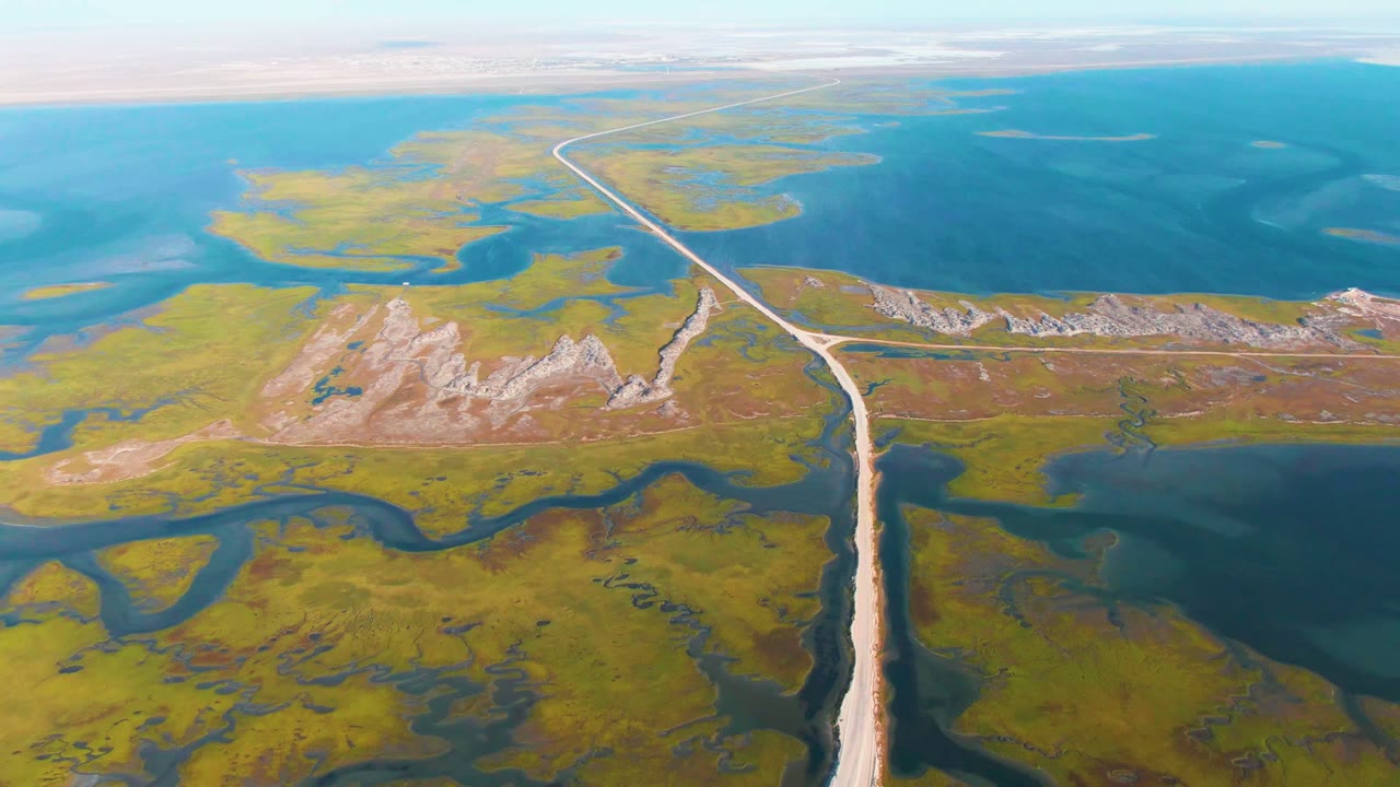 High aerial view near Guerrero Negro, Baja California Sur, showing a pale road running through green wetlands toward broad blue water, with low rocky hills and no visible people.