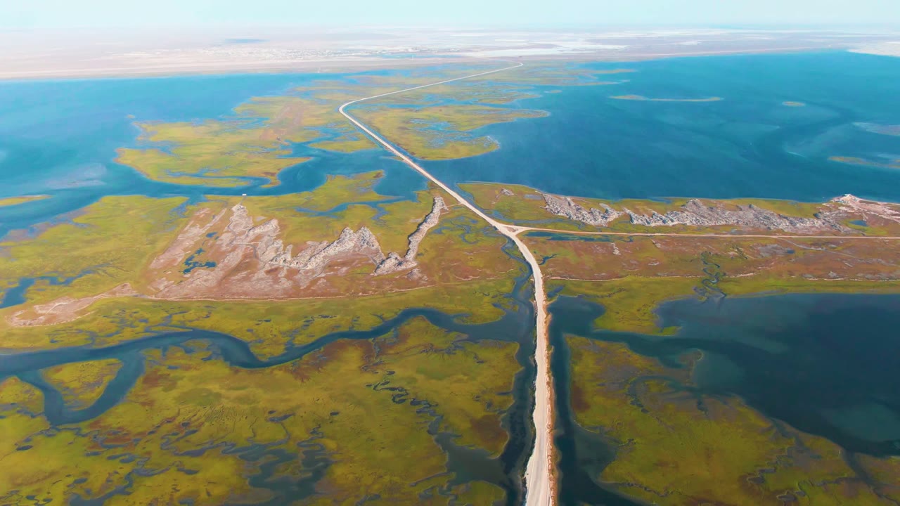 Wide aerial view in Guerrero Negro, Baja California Sur, with a pale road crossing between green wetlands and blue water while low rocky hills sit near the center, no visible people.