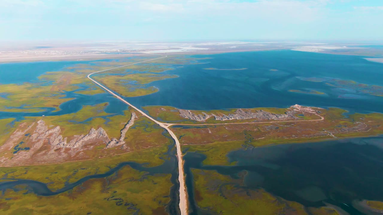 Aerial frame over Guerrero Negro, Baja California Sur, showing a pale dirt road running through green and brown marshland beside blue lakes, with low rocky ridges and no visible people.