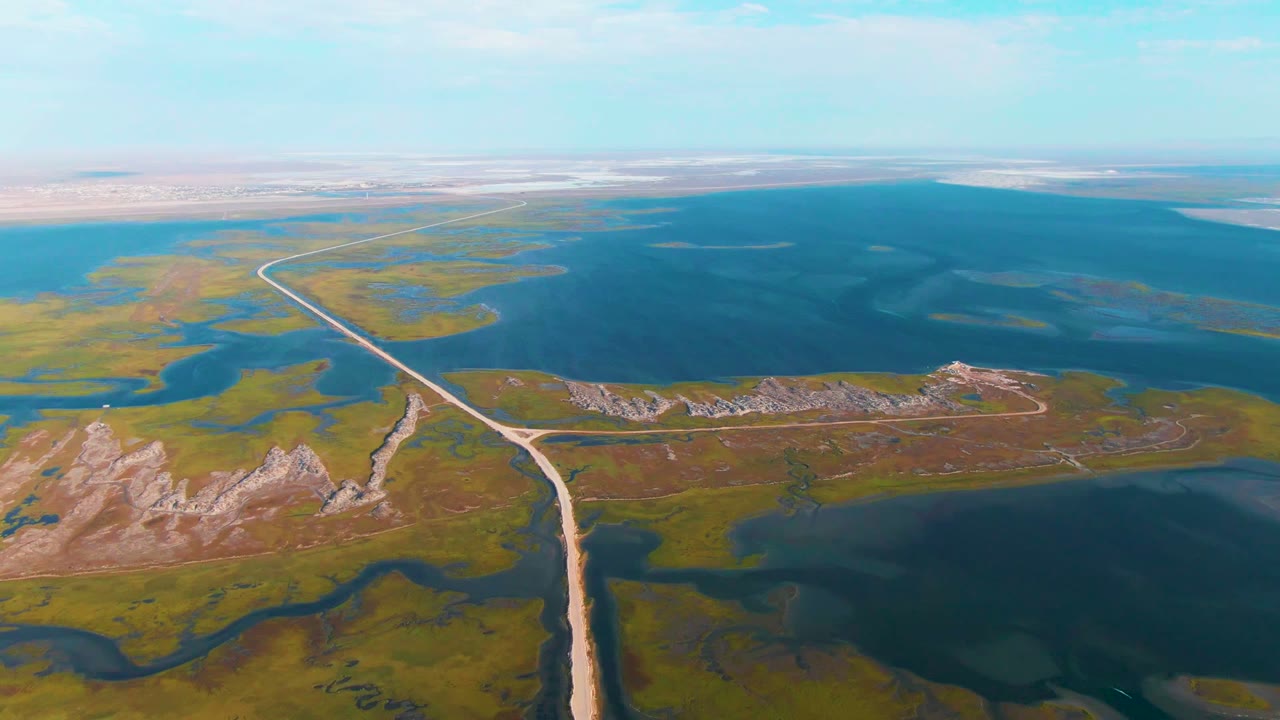 Drone-height view over Guerrero Negro showing the pale road splitting green marsh patches and blue water, with rocky foreground terrain and a long flat background, with no visible people.