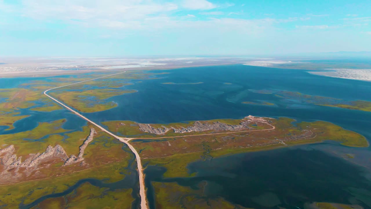 Aerial frame from Guerrero Negro, Baja California Sur showing a pale dirt road curving through green and brown wetlands beside dark blue water, with no visible people.