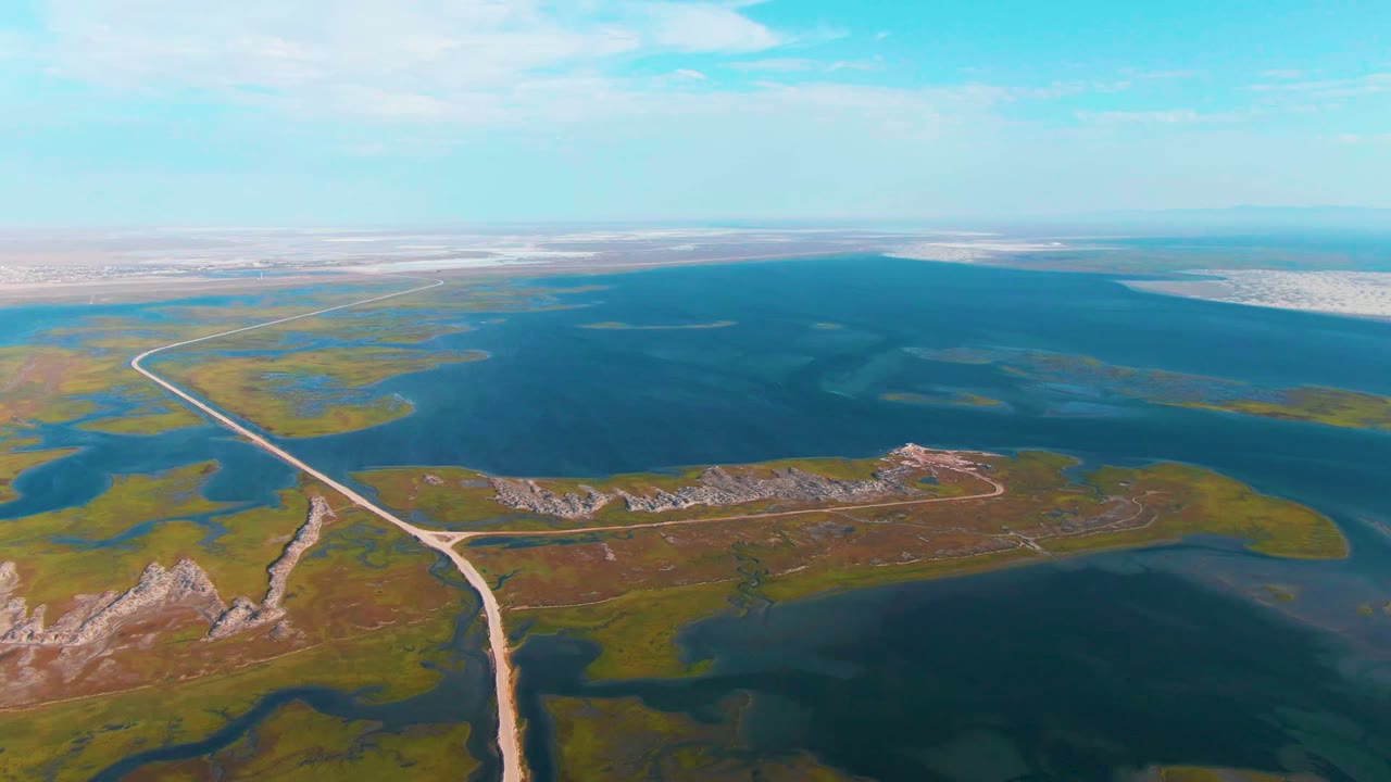 Aerial frame from Guerrero Negro, Baja California Sur showing the road bending past low vegetation and rocky ground toward blue water and pale distant flats, with no visible people.
