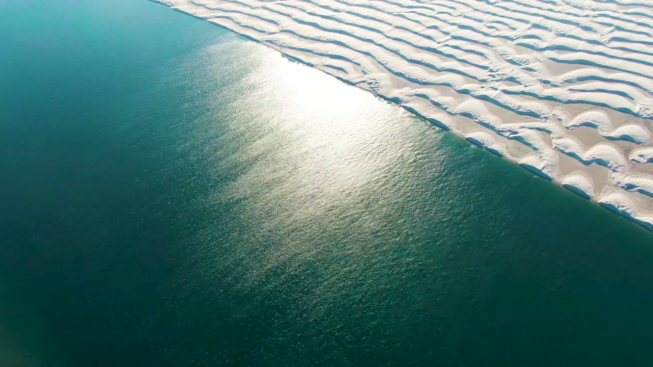 Opening aerial frame in Guerrero Negro, Baja California Sur, with rippled white dunes at upper right, turquoise water at lower left, bright sun reflection near the shore, and no visible people.