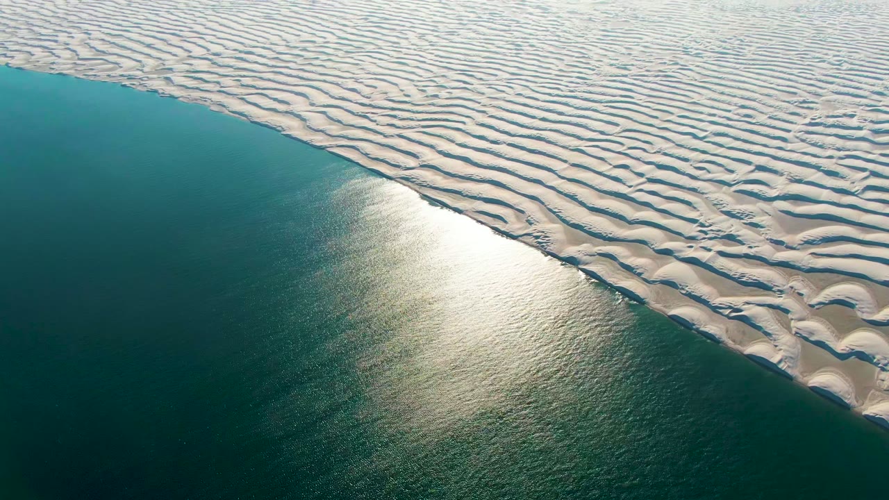 Aerial view in Guerrero Negro, Baja California Sur, showing pale rippled dunes meeting dark teal ocean water with a bright reflection near the left side and no visible people.