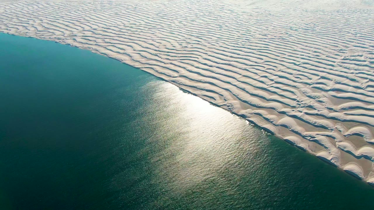 Bright reflection on calm ocean beside patterned sand ridges.