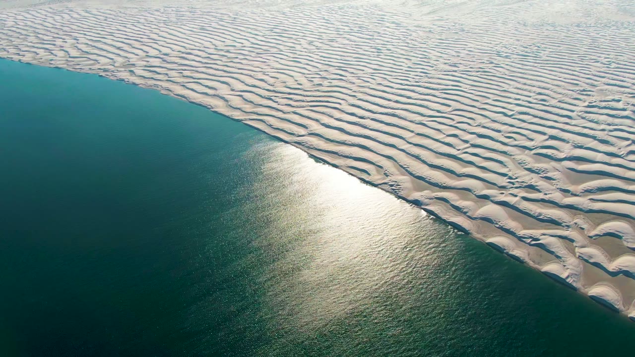 Oblique aerial frame over Guerrero Negro where white sand ripples run across the upper right and blue ocean fills the lower left, with no visible people.