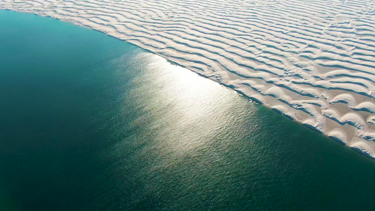 At 0:02, the aerial view over Guerrero Negro shows teal ocean in the lower frame and dense wavy white dune patterns across the upper frame, with no visible people.