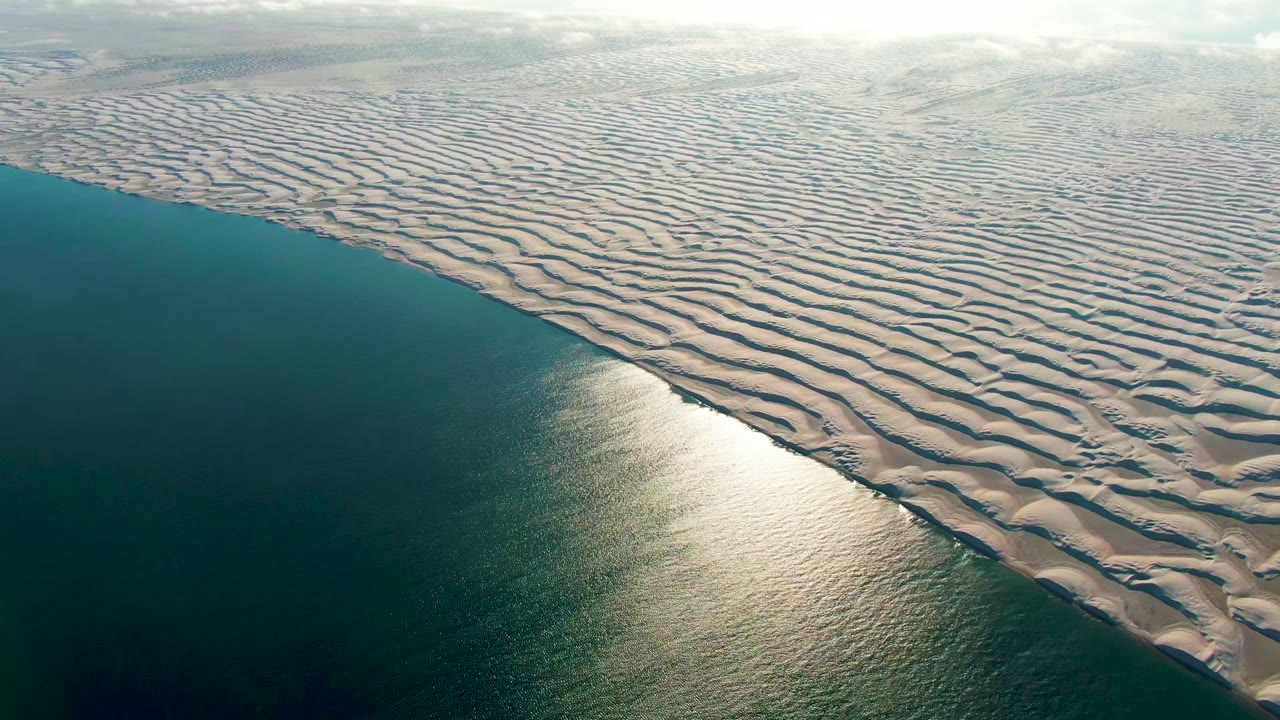Aerial frame from Guerrero Negro, Baja California Sur showing teal ocean on the left, beige rippled dunes on the right, bright water glare near the lower center, and no visible people.