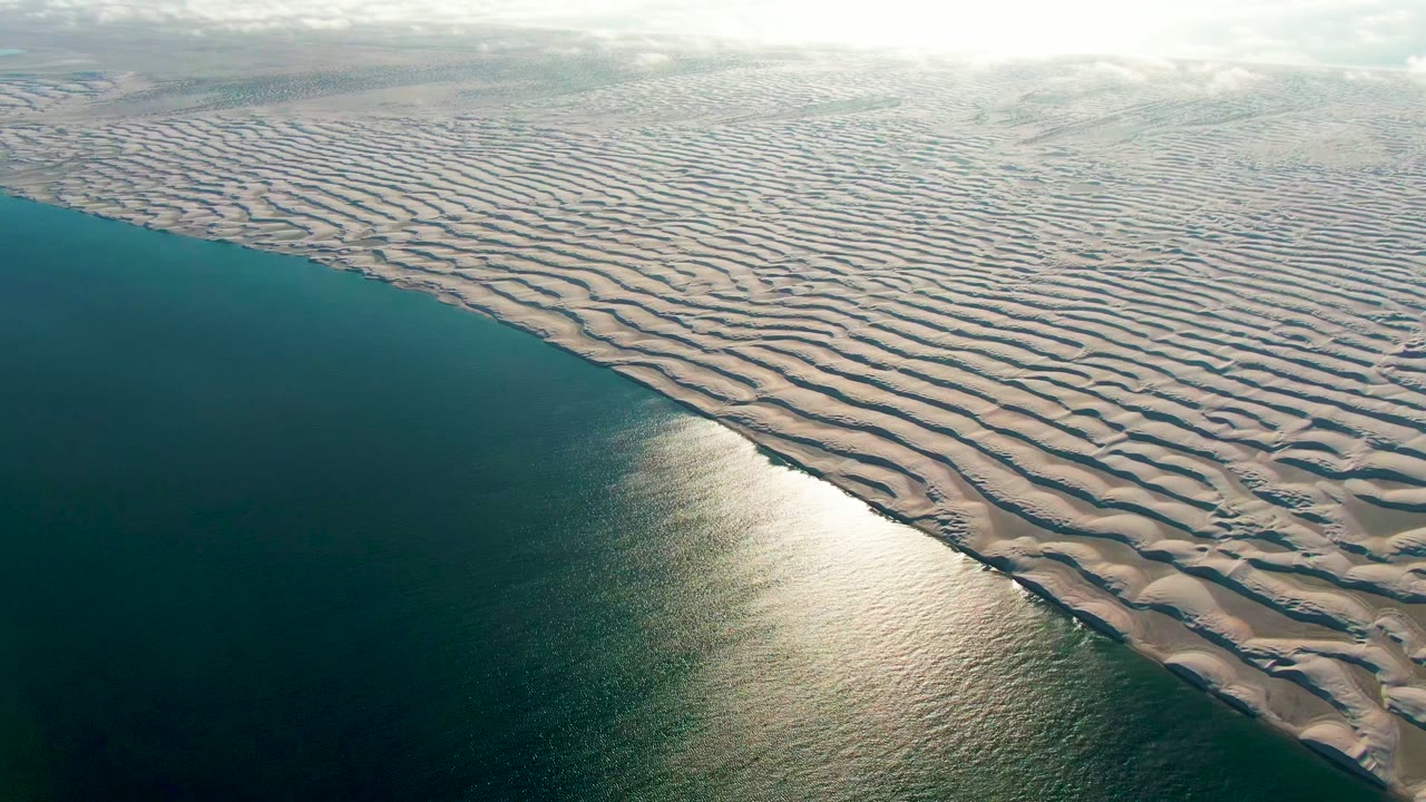 Calm ocean borders repeating rippled dunes in an aerial coastal view.