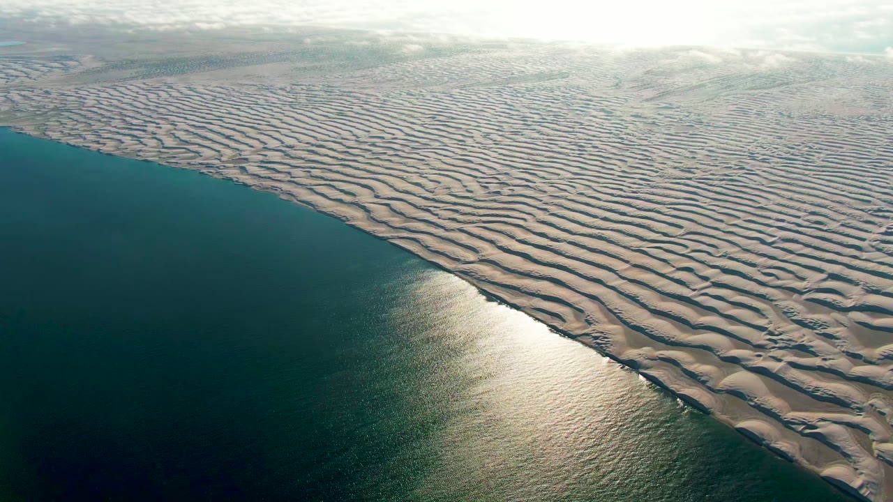 High aerial frame in Guerrero Negro, Baja California Sur with blue-green water occupying the lower left, a bright reflection near the shore, broad rippled sand patterns to the right, and no visible people.