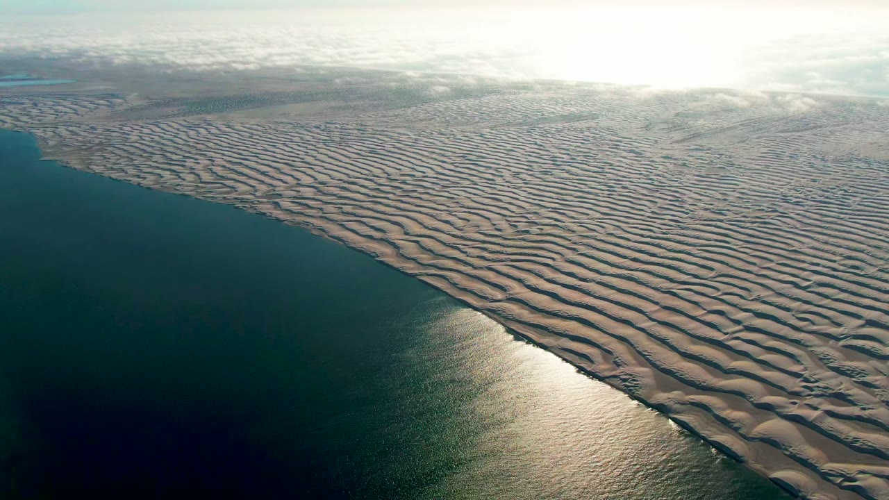 Opening aerial frame over Guerrero Negro, Baja California Sur, with dark teal ocean on the left, pale wavy sand ridges on the right, bright sky glare, and no visible people.