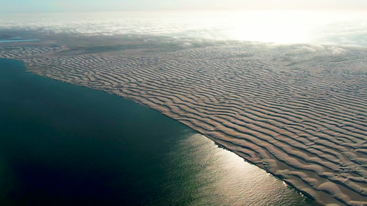 Wide aerial frame over Guerrero Negro, Baja California Sur, with ribbed pale sand filling the right side, dark ocean filling the left side, a bright reflection near the lower shore, and no visible people.