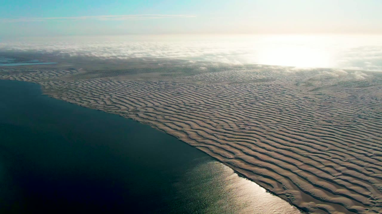 A wide aerial frame over Guerrero Negro, Baja California Sur, Mexico shows dark water along rippled wet sand, bright sun reflection near the horizon, pale clouds, and no visible people.