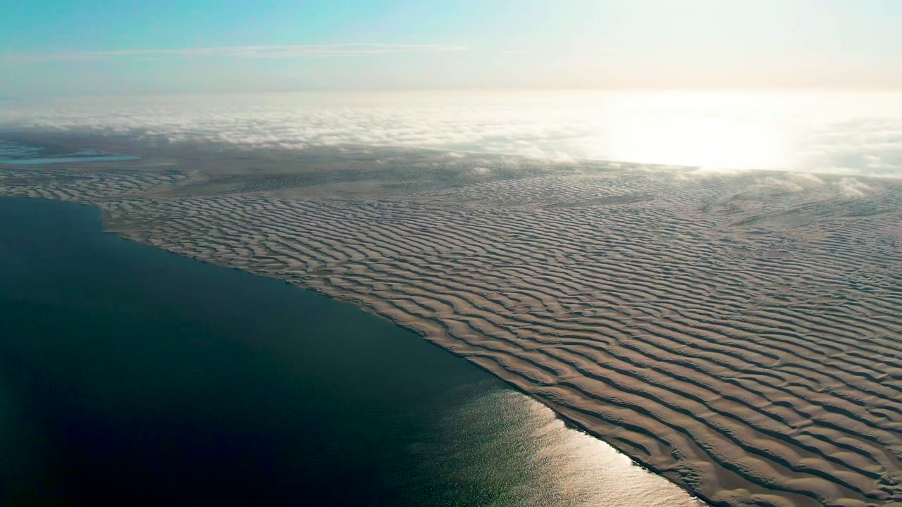 Elevated shoreline view with rippled sand bars and bright light on distant water.