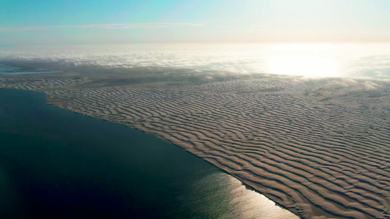 At 0:02, an elevated view over Guerrero Negro shows a broad field of parallel sand ripples meeting dark coastal water, with the brightest reflection concentrated near the upper right and no visible people.
