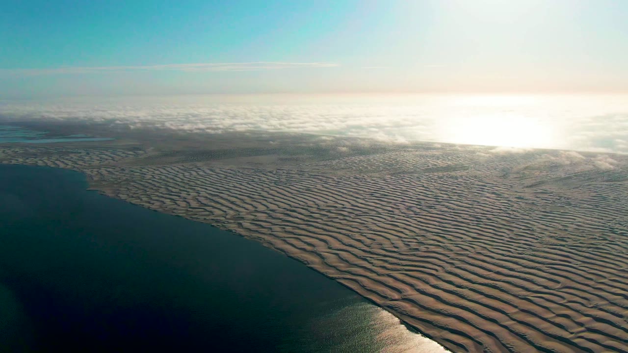 Aerial view near Guerrero Negro, Baja California Sur, with dark ocean on the left, rippled sand filling the foreground, bright sun glare on the right horizon, and no visible people.