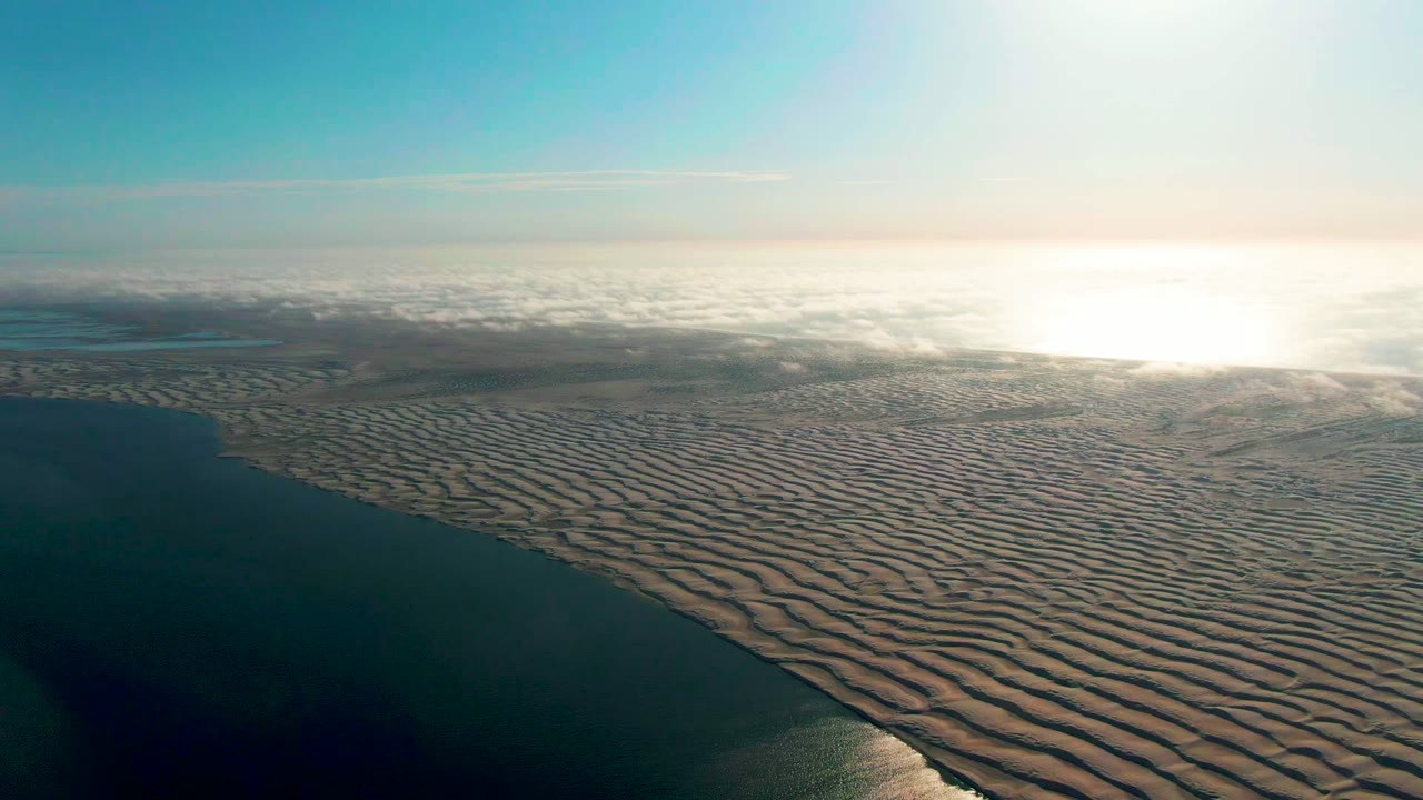 Aerial view near Guerrero Negro, Baja California Sur, with dark water at left, broad rippled sand across the lower frame, hazy cloud bands near the horizon, and no visible people.