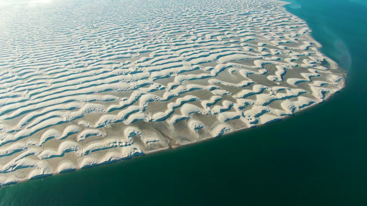 Aerial frame over Guerrero Negro, Baja California Sur showing pale wavy dunes curving into dark green-blue shoreline water, with no visible people.