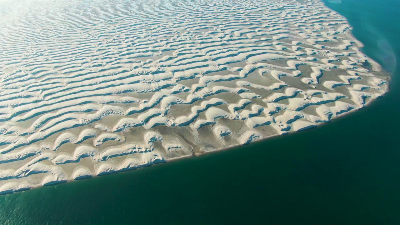 Oblique aerial frame over Guerrero Negro, Baja California Sur showing white and beige dune patterns ending at teal-green water along the right and lower edges, with no visible people.