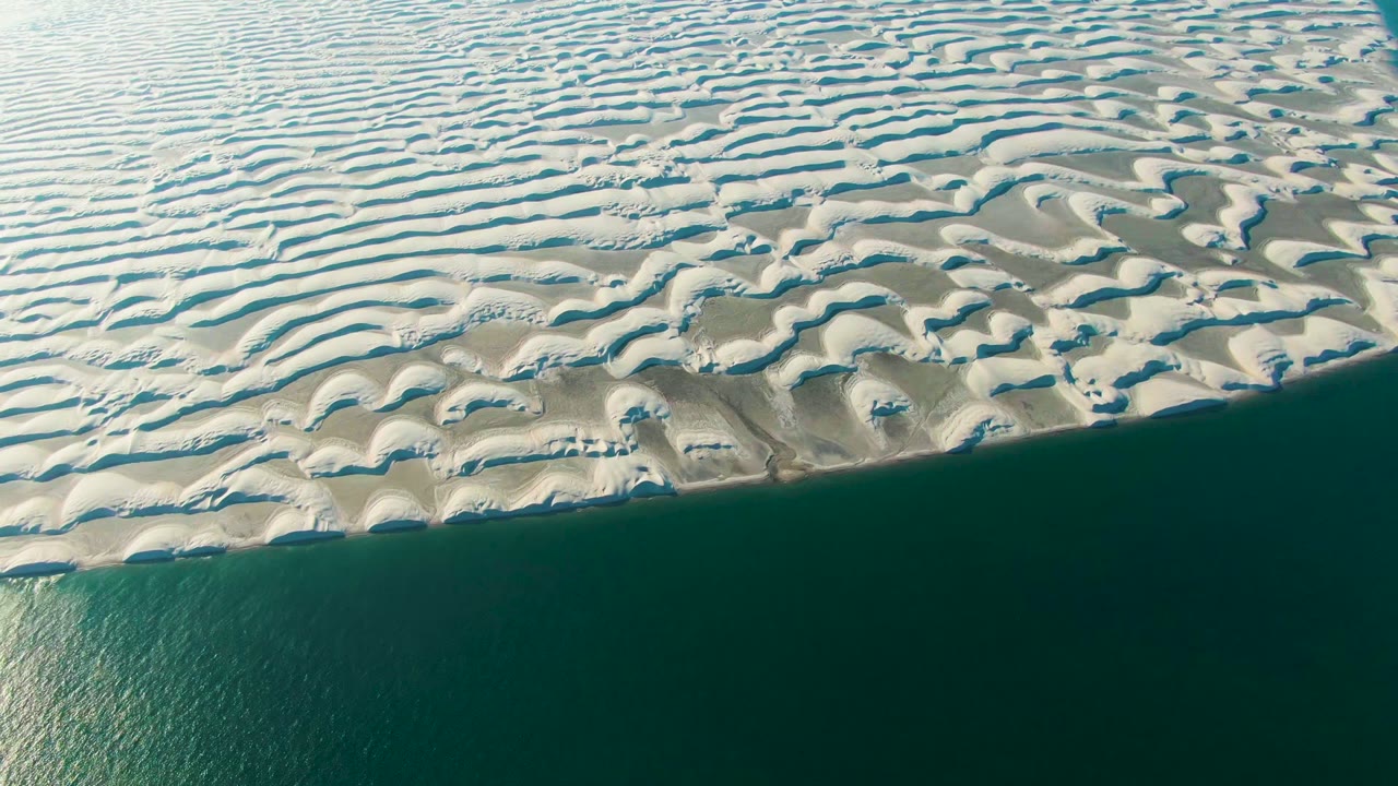 Aerial frame in Guerrero Negro, Baja California Sur, showing pale patterned dunes above dark teal ocean water with no visible people.