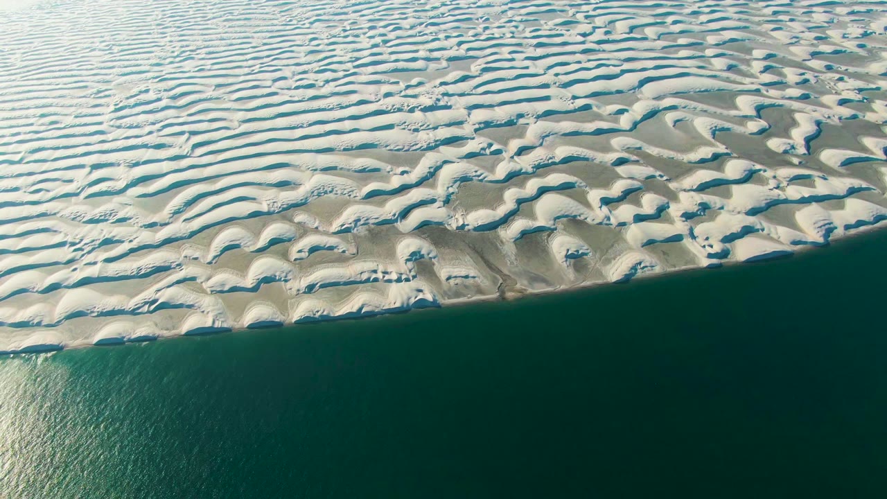 Patterned sand dunes meet calm blue-green ocean along a straight shoreline.