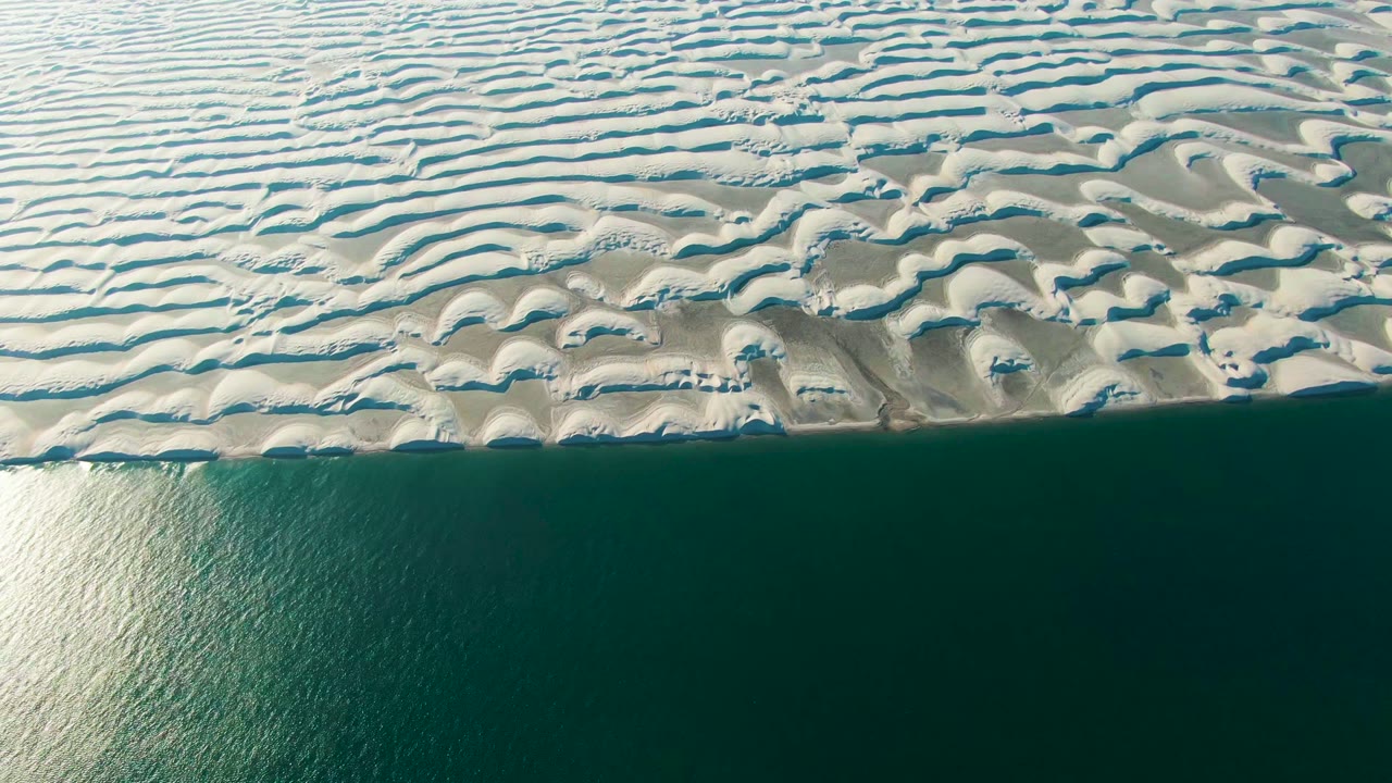 Overhead view in Guerrero Negro, Baja California Sur, with pale ribbed dunes across the top edge and dark blue-green ocean below, no visible people.
