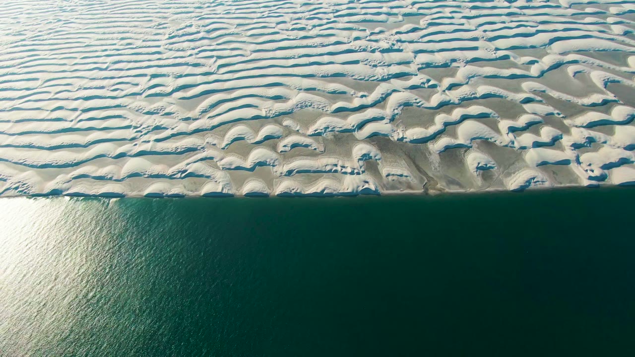 Overhead Guerrero Negro coastline with pale dune ripples above a narrow shore edge and calm blue-green ocean occupying the lower half, no visible people.