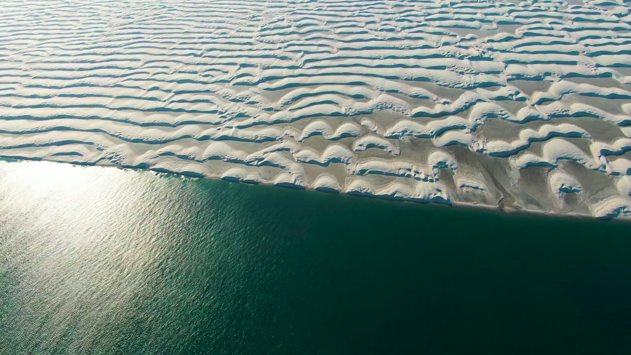 Aerial frame from Guerrero Negro, Baja California Sur, Mexico showing pale rippled sand dunes above calm blue-green water with bright surface reflection and no visible people.