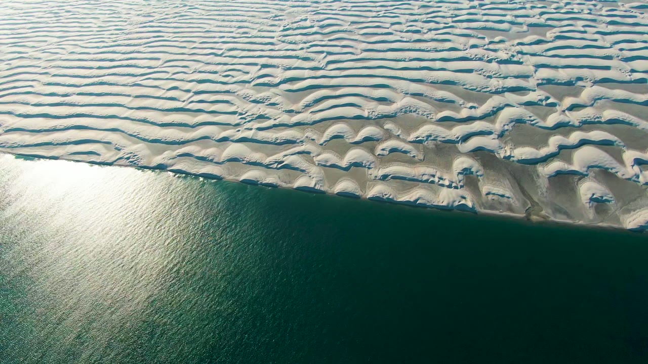 Rippled sand formations border dark calm water from an overhead view.