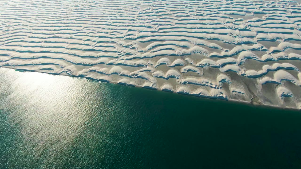 Aerial frame over Guerrero Negro showing pale dunes with shadowed channels meeting blue-green water, where the reflection sits near the lower left and no people are visible.