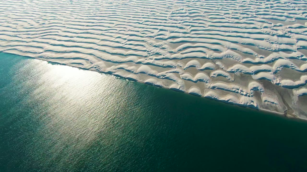 Aerial view in Guerrero Negro, Mexico, with white rippled dunes across the upper frame, green-blue ocean below, a bright water reflection near the left side, and no visible people.