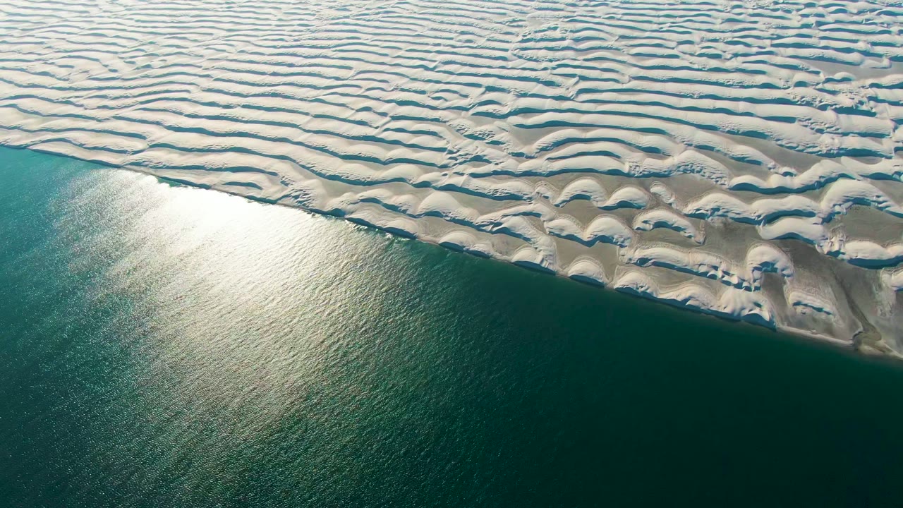Sunlit ocean edge below white patterned dunes.